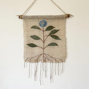 May include: A wall hanging made of light beige fabric featuring an embroidered blue flower with green leaves and brown roots. The fabric is suspended from a wooden dowel with a twine string, and loose brown threads hang from the bottom edge.