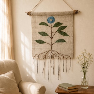 May include: A rectangular beige linen wall hanging featuring a hand-stitched botanical design. The artwork displays a single blue flower at the top, green leaves along a central stem, and exposed brown roots extending into long hanging threads.