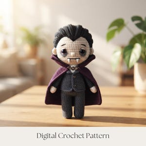 May include: A crocheted vampire doll featuring black hair, pale skin, and dark eyes. The figure wears a dark gray suit with two buttons and a deep purple cape with a small bat-shaped brooch at the collar. Two white fangs protrude from the mouth. Digital Crochet Pattern.