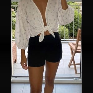 Puede incluir: Una blusa blanca de manga larga con un delicado estampado floral azul, con un escote en V profundo y un cierre de lazo frontal en la cintura, combinada con pantalones cortos negros lisos. La blusa tiene mangas voluminosas con puños fruncidos.