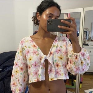 Puede incluir: Una blusa corta blanca con un estampado floral estilo acuarela en tonos rosa, amarillo y verde. La prenda tiene mangas largas y voluminosas con puños elásticos y un cierre de lazo frontal en el escote en V, creando una abertura delantera.