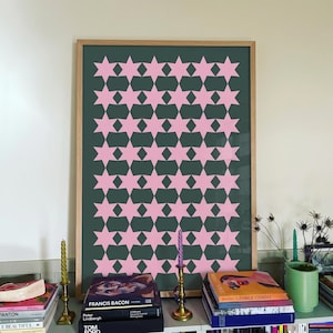 Può includere: Una stampa artistica da parete rettangolare in una cornice di legno chiaro con un motivo ripetuto di stelle a sei punte rosa chiaro su uno sfondo verde bosco scuro. Le stelle sono disposte in una griglia, creando un design geometrico.