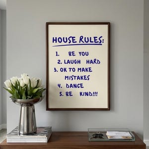 Può includere: Una stampa artistica incorniciata con sfondo color crema e testo scritto a mano in blu scuro che recita: HOUSE RULES: 1. BE YOU 2. LAUGH HARD 3. OK TO MAKE MISTAKES 4. DANCE 5. BE KIND!!! La stampa è racchiusa in una semplice cornice in legno marrone scuro.