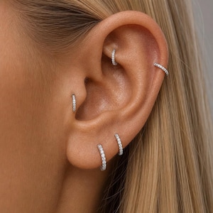 Può includere: Un set di quattro orecchini a cerchio color argento con piccole pietre chiare e scintillanti incastonate lungo il bordo esterno. Gli orecchini sono indossati in diverse posizioni sull'orecchio, tra cui lobo, trago, elice e rook.