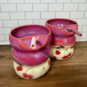 May include: Two ceramic tumblers with a rounded, bulbous shape. The upper portion features a vibrant, mottled magenta and purple glaze, while the lower section is cream-colored with hand-painted red cherry motifs. Each cup includes a straw.