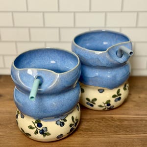 May include: Two ceramic tumblers with a rounded, stacked shape. The top half is glazed in a solid blue, while the bottom half is cream-colored with a hand-painted pattern of blue blueberries and green leaves. Each tumbler includes a light blue straw.