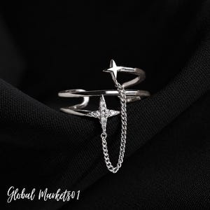 May include: A silver-toned open ring featuring two bands connected by a delicate chain. One band is topped with a four-pointed star, while the other features a larger, pavé-set four-pointed star charm that hangs from the chain, creating a layered look.