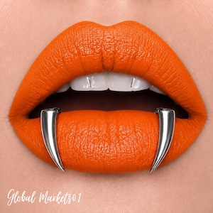 May include: A pair of silver-toned metal fangs positioned over the lower lip, contrasting with vibrant matte orange lipstick. The fangs are smooth, curved, and pointed, resting against the skin. The text Global Markets01 appears in white.