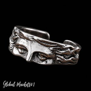 Puede incluir: Un anillo de banda abierta hecho de metal plateado, con un relieve detallado de un rostro con los ojos cerrados y cabello fluido. La superficie tiene un acabado pulido en las áreas elevadas y una textura oscura y oxidada en las partes hundidas.