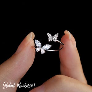 May include: A silver-toned open ring featuring two butterfly motifs. One butterfly is larger and adorned with marquise-cut clear stones, while the smaller butterfly is encrusted with tiny round clear stones. The band is thin and polished silver.