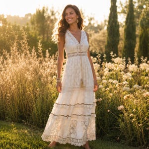 Peut inclure: Une robe longue blanche sans manches avec un décolleté en V, des détails en dentelle complexe, une taille ajustée avec une bande décorative et une jupe à étages avec une bordure en dentelle festonnée, ornée d'un long cordon à pompon.