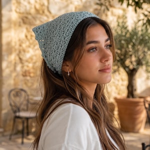 May include: A light blue triangular headscarf crafted with a textured crochet stitch pattern. The accessory features a delicate openwork design and is tied securely at the back of the head, covering the crown with a soft, matte yarn finish.