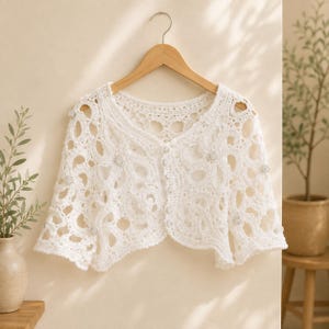 May include: A white crochet bolero cardigan featuring an intricate openwork pattern with circular motifs. The garment has short sleeves, a rounded neckline, and a front button closure with small, round, textured buttons integrated into the design.