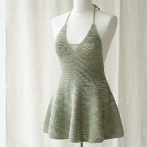 Könnte beinhalten: Ein salbeigrünes Häkel-Halterneck-Kleid mit tiefem V-Ausschnitt und einer Schnürung in der Mitte der Brust. Das Kleidungsstück hat eine ausgestellte Rock-Silhouette und dünne Träger, die im Nacken gebunden werden, gefertigt aus strukturiertem Garn.
