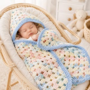 May include: A hooded baby blanket featuring a pastel granny square crochet pattern in shades of cream, pink, yellow, and light blue. The blanket has a solid light blue crocheted border along the edges and hood, wrapping around a sleeping infant.