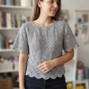 May include: A light gray short-sleeved top featuring an intricate openwork lace pattern throughout the fabric. The garment has a round neckline and a scalloped edge along the sleeves and the bottom hem, creating a delicate and textured appearance.