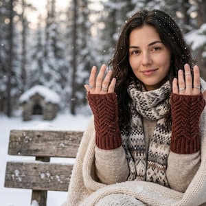 May include: A pair of dark brown fingerless gloves featuring a textured chevron knit pattern, worn with a beige sweater, a patterned scarf, and a large cream-colored knit blanket, with several silver rings on the fingers.