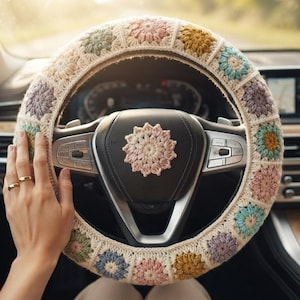 May include: A steering wheel cover featuring a crochet granny square pattern in cream, dusty pink, sage green, and mustard yellow. A matching circular crochet emblem is centered on the black steering wheel hub, surrounded by the textured yarn cover.