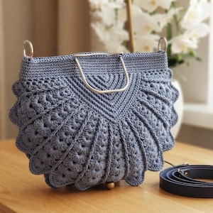 May include: A slate blue crochet handbag featuring a fan-shaped design with textured stitch patterns, a silver-toned metal semi-circular handle, and a detachable dark blue leather shoulder strap coiled on the surface next to the bag.