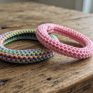 May include: Two circular bangles with a textured crochet finish. One bangle features a solid light pink yarn, while the other displays a multi-colored pattern of pastel yellow, light blue, lavender, and green yarn woven in a repeating horizontal stripe.