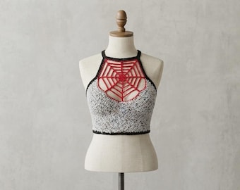 May include: A crochet crop top featuring a grey and black speckled yarn body with a black trim. The upper section displays a red spiderweb pattern integrated into the neckline, creating a halter-style design on a beige mannequin torso.