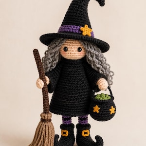 Pode incluir: Uma boneca bruxa de crochê com cabelo de lã cinza, vestindo um vestido preto, leggings listradas roxas e um chapéu pontudo preto com uma faixa roxa e uma estrela amarela. A boneca segura uma vassoura de lã marrom e um caldeirão preto.