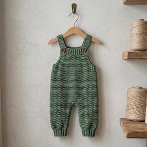 May include: A sage green crocheted romper featuring a ribbed neckline and leg cuffs, two shoulder straps secured with small brown buttons, and a textured stitch pattern. The garment is displayed on a wooden hanger against a neutral wall.