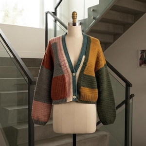 May include: A long-sleeved, V-neck cardigan featuring a color-blocked design with panels of mustard yellow, olive green, dusty pink, slate gray, and teal. The knit garment has a button-front closure with two visible light-colored buttons.