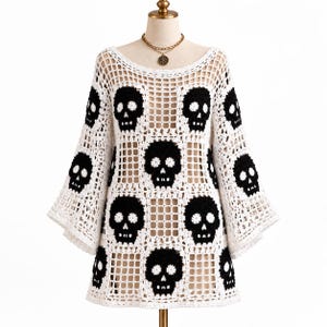 May include: A white crochet top featuring a repeating black skull pattern throughout the garment. The top has a wide, open-knit grid construction, long flared sleeves, and a round neckline, displayed on a mannequin with a gold chain necklace.
