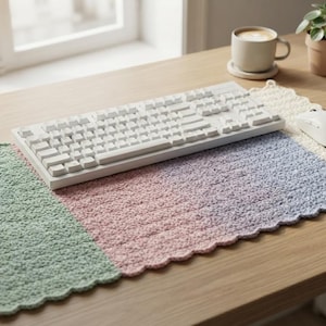 May include: A rectangular crochet desk mat featuring a color-blocked design with sections of sage green, soft pink, lavender, and cream. The mat has a scalloped edge and is positioned under a white computer keyboard and a white computer mouse.