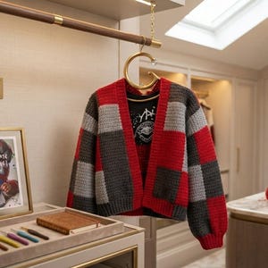 May include: A crochet cardigan featuring a red, gray, and black checkered pattern, hanging on a gold G-shaped hanger. Below, a framed print displays the text The (Almost) Ultimate Patchwork Cardigan, alongside a tray with crochet hooks.