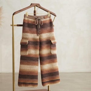 Könnte beinhalten: Häkel-Cargohose mit horizontalem Streifenmuster in Braun-, Beige- und Cremetönen. Die Hose verfügt über einen Kordelzug in der Taille, weit geschnittene Beine und zwei große, rechteckige Cargotaschen an den äußeren Oberschenkeln.