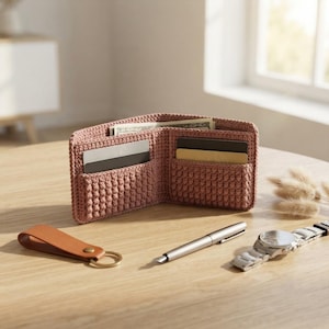 May include: A dusty rose crocheted bifold wallet with textured stitching, containing paper currency and two blank cards in its slots. Beside it lies a tan leather key fob, a silver metal fountain pen, and a silver-toned analog wristwatch.