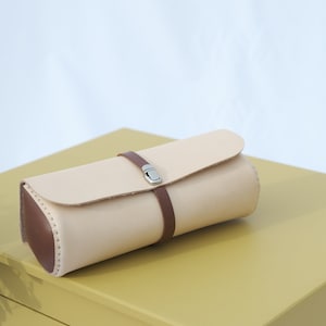 May include: A light brown leather pencil case with a brown leather strap and a silver buckle. The case is sitting on a yellow wooden box with three drawers.