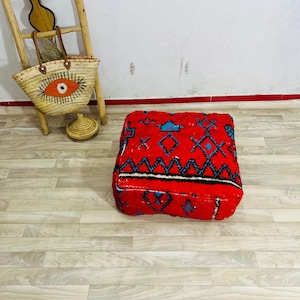 May include: A square floor pouf featuring a vibrant red base with intricate blue and black geometric patterns. The textile surface displays a textured, woven appearance with repeating diamond and zigzag motifs across the top and side panels.