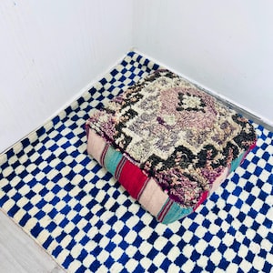 May include: A square floor pouf featuring a thick, shaggy top surface with a muted purple, cream, and dark brown geometric pattern. The sides of the pouf are constructed from fabric panels in solid stripes of bright red, teal, and light pink.