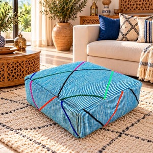 May include: A square floor cushion featuring a textured light blue fabric with a fine white striped pattern. The surface is decorated with intersecting lines in vibrant colors including black, red, green, blue, and orange, creating a geometric design.