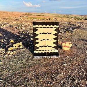 May include: A rectangular rug featuring a cream-colored center with black geometric zig-zag patterns and black borders with white wavy lines, finished with white fringe. Beside it sits a woven straw basket with the red text Marrakech.