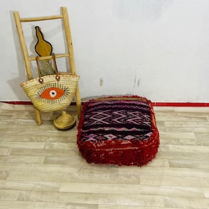 May include: A rectangular floor cushion featuring a deep purple and red woven pattern with fringed edges, accompanied by a straw tote bag with a prominent orange and black eye design, a small lidded straw basket, and a light wood ladder.