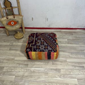 May include: A square floor pouf featuring a dark purple top panel with intricate white and multi-colored geometric patterns, and side panels with vertical stripes in shades of orange, yellow, pink, and black, constructed from woven textile.
