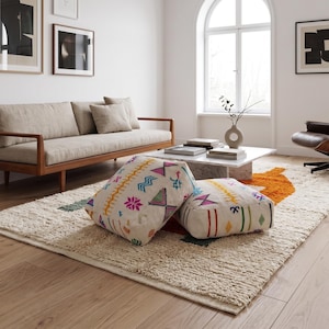 May include: Two square floor cushions featuring an off-white fabric base with colorful embroidered geometric patterns in shades of pink, orange, green, and blue. The cushions are placed on a textured cream-colored area rug with orange accents.