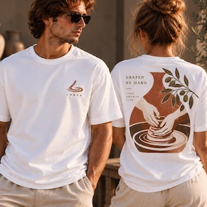 Könnte beinhalten: Ein weißes kurzärmeliges T-Shirt mit einem kleinen braunen Logo auf der Brust und dem Schriftzug LORYX. Die Rückseite zeigt eine Grafik von Händen beim Töpfern, den Text SHAPED BY HAND und LORYX CERAMICS CLUB sowie einen Olivenzweig.
