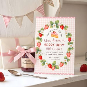 Puede incluir: Una tarjeta de invitaci&oacute;n de cumplea&ntilde;os rectangular con un borde de cuadros vichy rosa y una ilustraci&oacute;n de una corona de fresas. El texto dice: You are invited to Charlotte's Berry First Birthday, 2pm, 5th August 2026, at Open House Dining, RSVP to Julia at 412-456-7890.