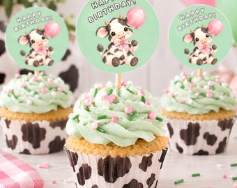 Könnte beinhalten: Drei Cupcakes in Förmchen mit Kuhfleckenmuster, garniert mit hellgrünem Zuckerguss, rosa und weißen Streuseln sowie runden Aufsteckern, die eine Comic-Kuh mit einem rosa Luftballon und dem Schriftzug HAPPY BIRTHDAY zeigen.
