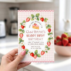 Puede incluir: Una tarjeta de invitaci&oacute;n rectangular con fondo de cuadros vichy rosa claro y borde festoneado blanco. Una corona de fresas y flores blancas rodea el texto: YOU ARE INVITED TO, Charlotte's BERRY FIRST BIRTHDAY, 2PM, 5TH AUGUST 2026, AT OPEN HOUSE DINING, RSVP TO JULIA AT +123-456-7890.