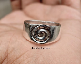 Peut inclure: Une bague en argent poli présentant un motif en spirale en relief sur la bande avant. La surface métallique est lisse et réfléchissante, mettant en valeur un motif tourbillonnant circulaire. Le texte RaiSilverJewelers est imprimé en noir sous la bague.