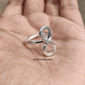 Peut inclure: Une bague en argent poli pr&eacute;sentant un motif de symbole infini vertical. L'anneau est lisse et circulaire, se connectant &agrave; la forme en huit qui s'&eacute;tend vers le haut. Le texte RaiSilverJewelers est imprim&eacute; en noir pr&egrave;s de la base de la bague.