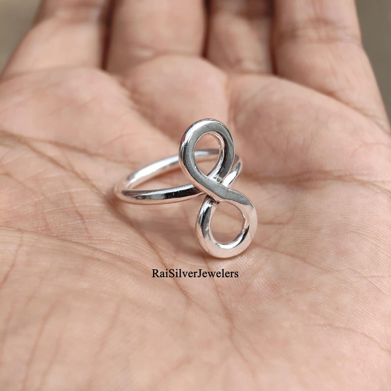 Peut inclure: Une bague en argent poli pr&eacute;sentant un motif de symbole infini vertical. L'anneau est lisse et circulaire, se connectant &agrave; la forme en huit qui s'&eacute;tend vers le haut. Le texte RaiSilverJewelers est imprim&eacute; en noir pr&egrave;s de la base de la bague.