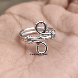 Puede incluir: Un anillo de plata con un diseño de doble banda y dos bucles de alambre envuelto en el centro. El metal tiene un acabado pulido y el texto RaiSilverJewelers está impreso en negro cerca de la base del anillo. La estructura consta de dos bandas paralelas que convergen en bucles decorativos de alambre trenzado.