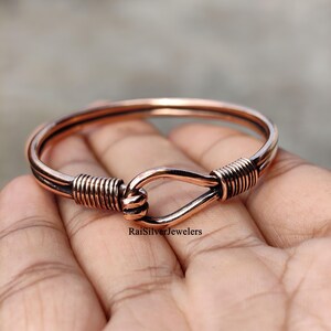 Può includere: Un bracciale rigido in metallo color rame caratterizzato da una chiusura a gancio con dettagli in filo avvolto alle estremità. Il bracciale ha una finitura liscia e lucida e include il testo RaiSilverJewelers stampato sulla superficie.